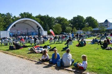 Ein Open-Air-Konzert im Park mit einer großen Menge an Menschen, die auf Stühlen sitzen oder auf dem Gras liegen, während eine Band auf der Bühne im Hintergrund spielt, umgeben von grünen Bäumen und einem blauen Himmel.