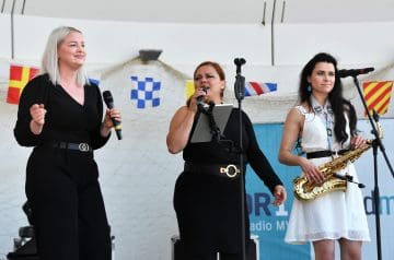 Drei Frauen stehen auf einer Bühne bei einem Musikauftritt. Die Frau auf der linken trägt schwarze Kleidung, hat blonde Haare und hält ein Mikrofon. Die mittlere Frau ist dunkelhaarig, trägt ebenfalls schwarze Kleidung, hält ein Mikrofon und hat eine Pult mit einem digitalen Tablet vor sich. Die Frau auf der rechten hat schwarze Haare, trägt eine weiße Kleidung mit einem schwarzen Gürtel und hält eine goldene Saxophon, während sie vor einem Mikrofon steht. Im Hintergrund sind Flaggen und ein Netz an der Wand sichtbar.