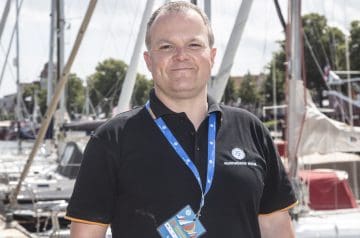 Mann mit kurzen grauen Haaren, trägt schwarzes Polo-Shirt mit Logo und Namensschild, steht vor einem Hafen mit Segelbooten.