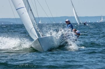 Segelboote auf dem Wasser bei regem Wind, eine Crew ist in einem auf Schräglage fahrenden Boot sichtbar, mit Wellen und weiteren Booten im Hintergrund.