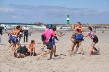 Menschen beim Strand-Flag Rugby, einige Frauen in rot-weiß karierten Trikots, einige Männer und Kinder, im Hintergrund der Ozean mit Wellen, ein Leuchtturm und eine Mole mit Vögeln am Himmel.