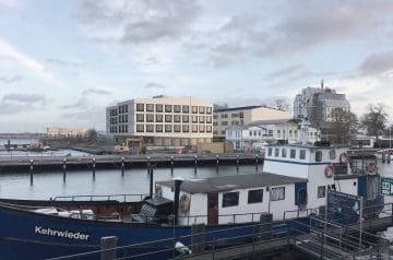 Ein Segelschiff namens Kehrwieder liegt im Hafen, im Hintergrund moderne Gebäude und einige Bauarbeiten, bei bewölktem Himmel.