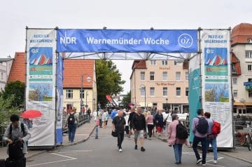 Straßenansicht eines Festes mit einem großen blauen Banner am Eingang, auf dem steht 'Warnemünder Woche', umgeben von mehreren Menschen, einige mit Rucksäcken, eine Person mit roter Tasche und eine Frau mit rotem Regenschirm, im Hintergrund historische Gebäuden, einige Bäume und Straßenlaternen.