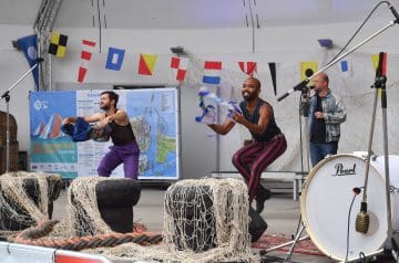 Drei männliche Performer auf einer Bühne, die in einem Raum mit Flaggen an der Wand und einem hohen Deckenhintergrund tanzen oder auftreten, während sie Tücher in ihren Händen halten und musizieren, mit Musikinstrumenten wie einer Trommel im Vordergrund.