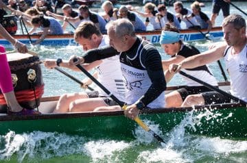 Mehrere Menschen in einem Wettkampfboot paddeln energisch durch das Wasser, mit einem Hintergrund von weiteren Paddlern, die sich an einem Rennen beteiligen.