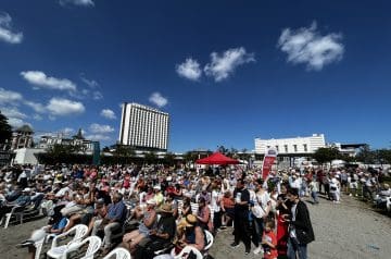 Ein großes öffentliches Event mit vielen Menschen, die im Freien sitzen und stehen, vor einem blauen Himmel mit wenigen Wolken, in einer Stadt mit modernen Gebäuden im Hintergrund.