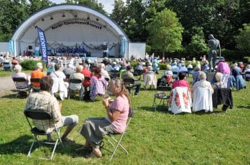 Ein Open-Air-Konzert im Freien mit vielen Zuschauern, die auf Stühlen sitzen und eine Band auf der Bühne im Hintergrund spielen, umgeben von Bäumen und einer Skulptur einer nackten Person.