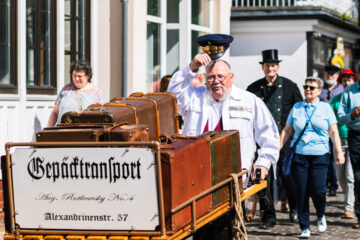 Ein älterer Mann in Weiß mit Brille und weißem Bart, der einen schwarzen Hut hebt, steht vor einem Wagen mit alten Koffern, während eine Gruppe von Menschen im Hintergrund bei einer Parade oder einem Fest unterwegs ist.