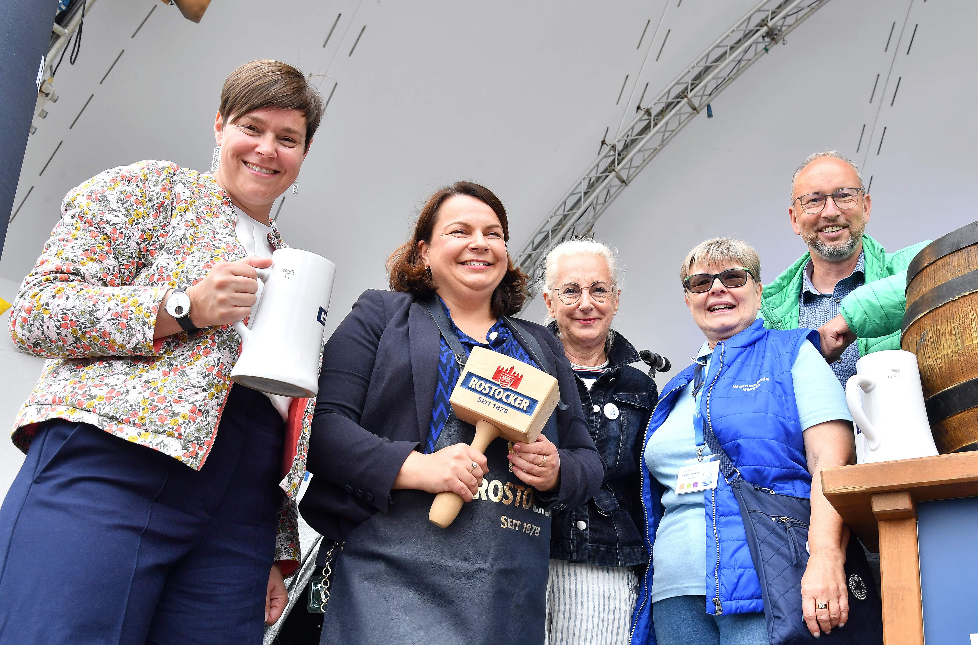 Gruppe von fünf Menschen, die auf einer Bühne stehen, einige halten Bierkrüge, und eine Frau hält ein Holzhammer mit einem Rostocker Logo drauf, lächeln in die Kamera.