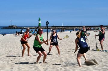 Sechs Frauen spielen Beach Lacrosse am Strand, mit einer Sandfläche, im Wasser und einem Leuchtturm im Hintergrund.