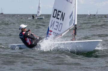 Seglerin in Wettkampfausrüstung auf einem kleinen Boot, das über das Wasser fährt, mit mehreren anderen Booten im Hintergrund bei einem Segelrennen.