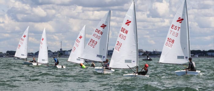 Segelboote mit weißen Segeln auf dem Wasser, einige mit roten Markierungen und Nummern, bei bewölktem Himmel in Küstenstadt im Hintergrund.