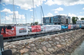 Blick auf einen Kai mit Segelbooten im Wasser, umgeben von einer Steinmauer, mit Werbebannern entlang des Ufers, moderne Gebäude im Hintergrund und ein blauer Himmel mit weißen Wolken.