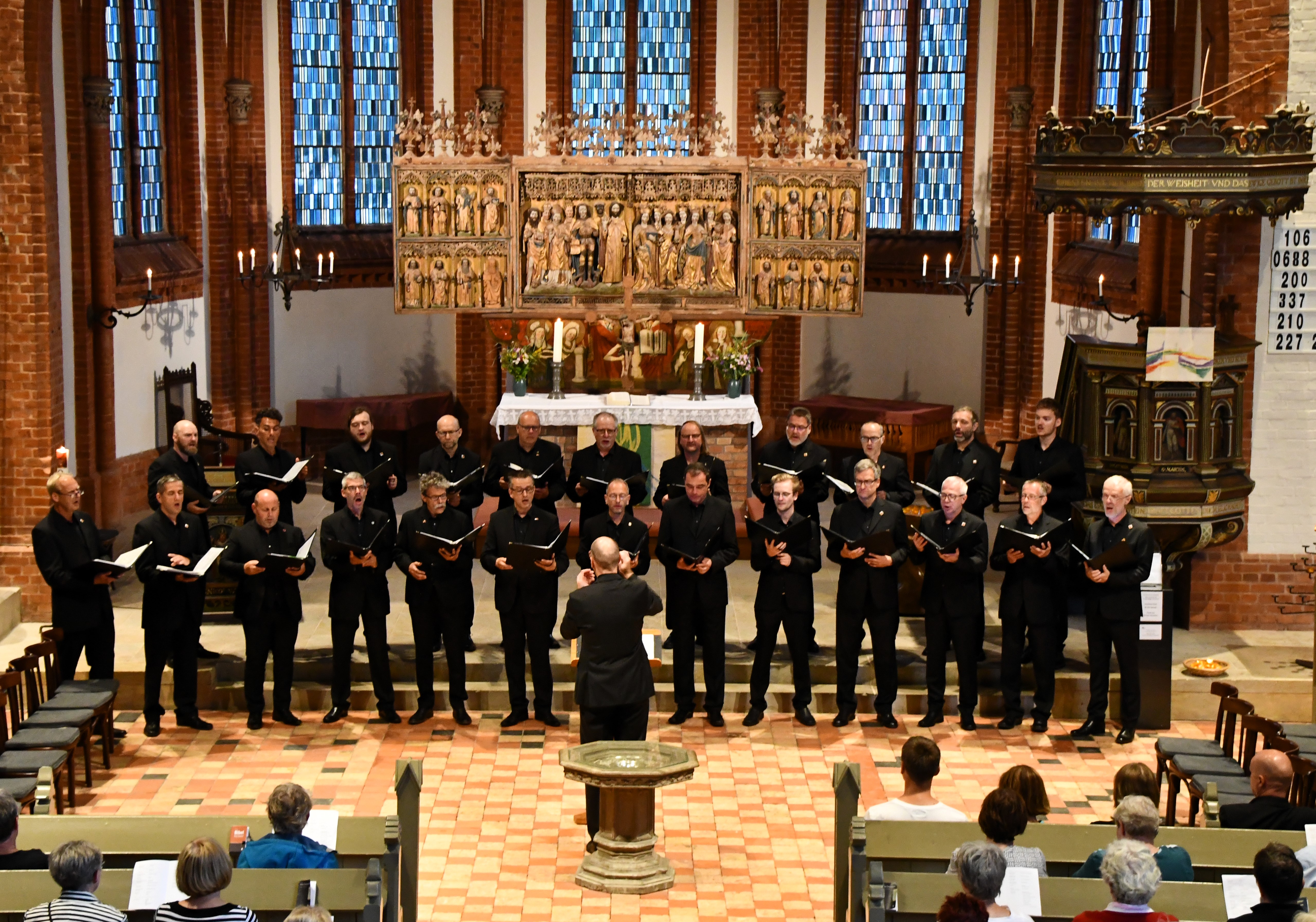 Chor von Männern in schwarzen Anzügen singt in einer Kirche vor einem Altar mit Blumen und Kerzen, im Hintergrund bunte Buntglasfenster und kunstvolle Holzdekor.