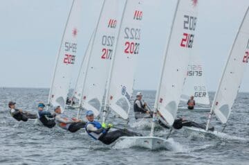 Mehrere Segelboote mit weißen Segeln, die auf dem Wasser bei einem Regatta-Wettbewerb segeln, mit jeweils mehreren Crewmitgliedern an Bord, einige mit Sonnenbrillen und Hüten, und das Meer im Hintergrund.