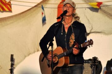 Ein älterer Mann mit langem, grauem Haar, der eine Mütze trägt, steht mit einer akustischen Gitarre vor einem Mikrofon und singt. Im Hintergrund sind bunte Fähnchen und ein Netz sichtbar.