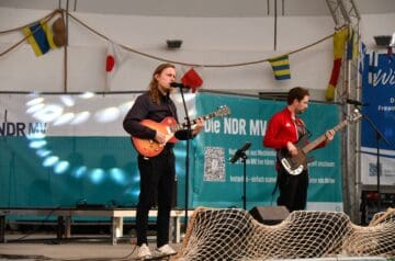 Zwei Musiker auf einer Bühne, einer spielt Gitarre und singt, der andere spielt Bass, im Hintergrund eine blaue Werbefläche mit Text und QR-Code, dekorative Flaggen hängen.