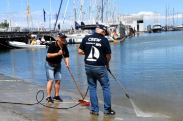 Zwei Personen reinigen einen Hafenbucht. Eine Frau mit schwarzen Polo-Shirt, Jeansshorts und Baseballkappe benutzt eine Bürste, während ein Mann im gleichen Polo-Shirt mit Crew-Aufdruck und Jeanshose mit einem Wasserschlauch Wasser aufspritzt.