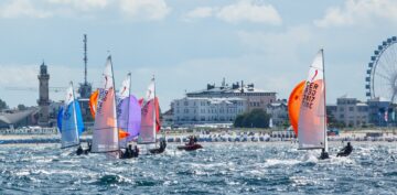 Segelboote auf dem Wasser bei einer Regatta, mit einem Stadtstrand im Hintergrund, einem Leuchtturm und einem Riesenrad, bei bewölktem Himmel.