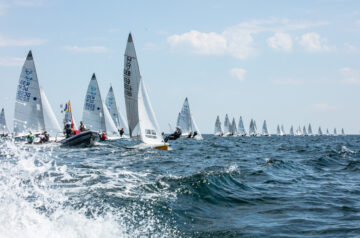 Segelboote auf dem Meer bei einer Regatta, mit einigen Crewmitgliedern in den Booten, blaue und graue Wolken am Himmel, leichte Wellen im Wasser.