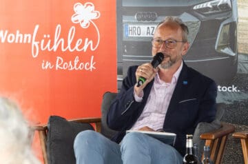 Ein Mann mit Brille, grauem Bart und grauem Haar, trägt einen dunkelblauen Anzug und ein helles Hemd, sitzt in einem Sessel mit grauem Kissen, hält ein Mikrofon in der Hand und spricht bei einer Veranstaltung, im Hintergrund ist ein Auto und eine rote Werbetafel mit weißen Schriftzug, die "Wohlfühlen in Rostock" zeigt.