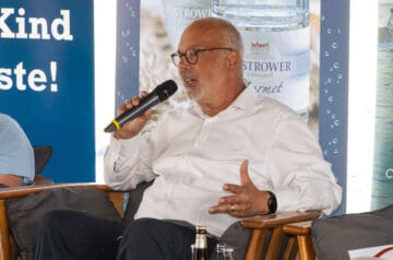 Ein älterer Mann mit Glatze, Brille und grauem Bart sitzt auf einem Stuhl, hält ein Mikrofon in der Hand und trägt ein weißes Hemd. Im Hintergrund sind Wasserflaschen und ein Banner mit Wasser- und Mineralwasserwerbung sichtbar.