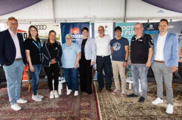 Eine Gruppe von elf Personen steht nebeneinander in einem Zelt, im Hintergrund sind Banner und Sessel sichtbar.