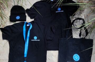 Verschiedene schwarze Kleidungsstücke und Accessoires mit dem Logo "Warnemünder Woche", darunter eine Mütze, ein T-Shirt, eine Latzhose, ein Rucksack und eine Lanyard, liegen auf Sand, umgeben von Gras.