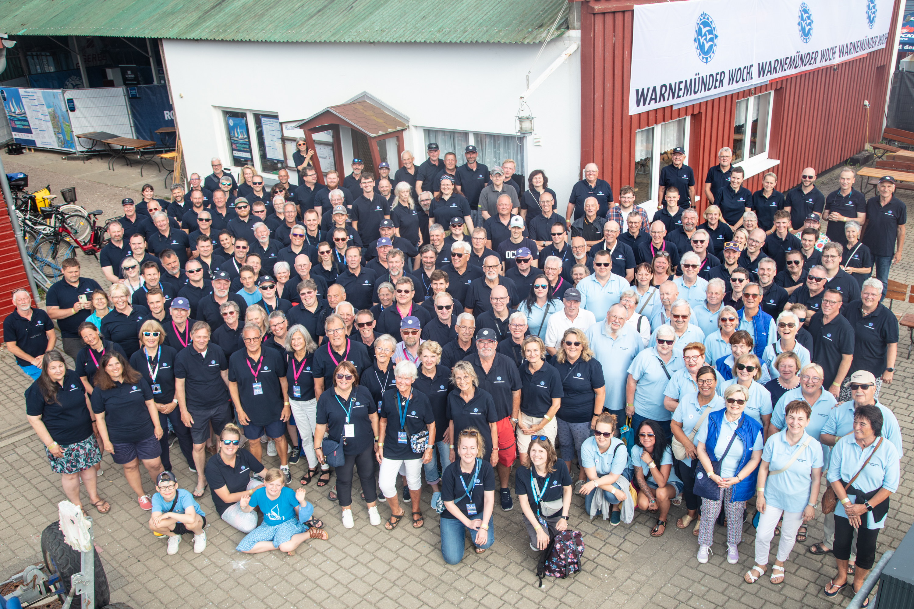 Gruppenfoto mit vielen Menschen vor einem Gebäude mit einem Banner, auf dem 'Warnemünder Woche' steht, alle tragen dunkelblaue Polohemden.